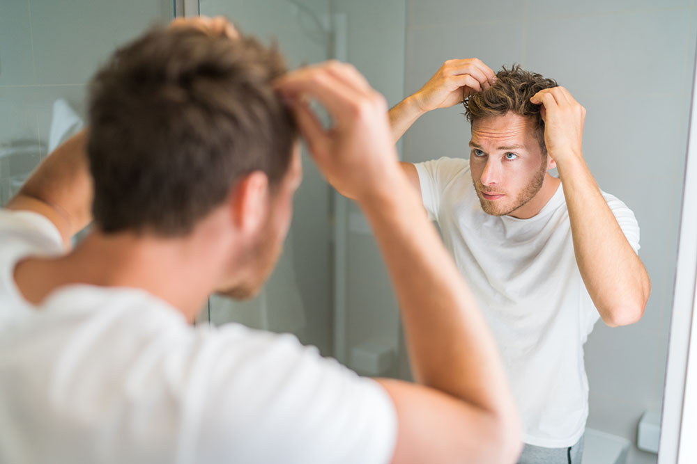 Hair loss man looking in bathroom mirror putting wax touching his hair styling or checking for hair loss problem. Male problem of losing hairs.